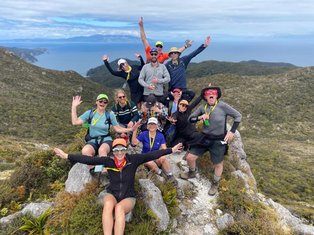 A group of hikers stand triumphantly atop the Te Ahumata/Whitecliffs summit on Aotea/Great Barrier Island, on a guided walk in New Zealand