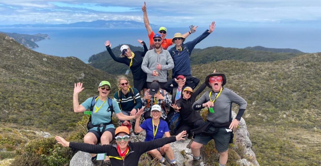 A group of hikers stand triumphantly atop the Te Ahumata/Whitecliffs summit on Aotea/Great Barrier Island, on a guided walk in New Zealand