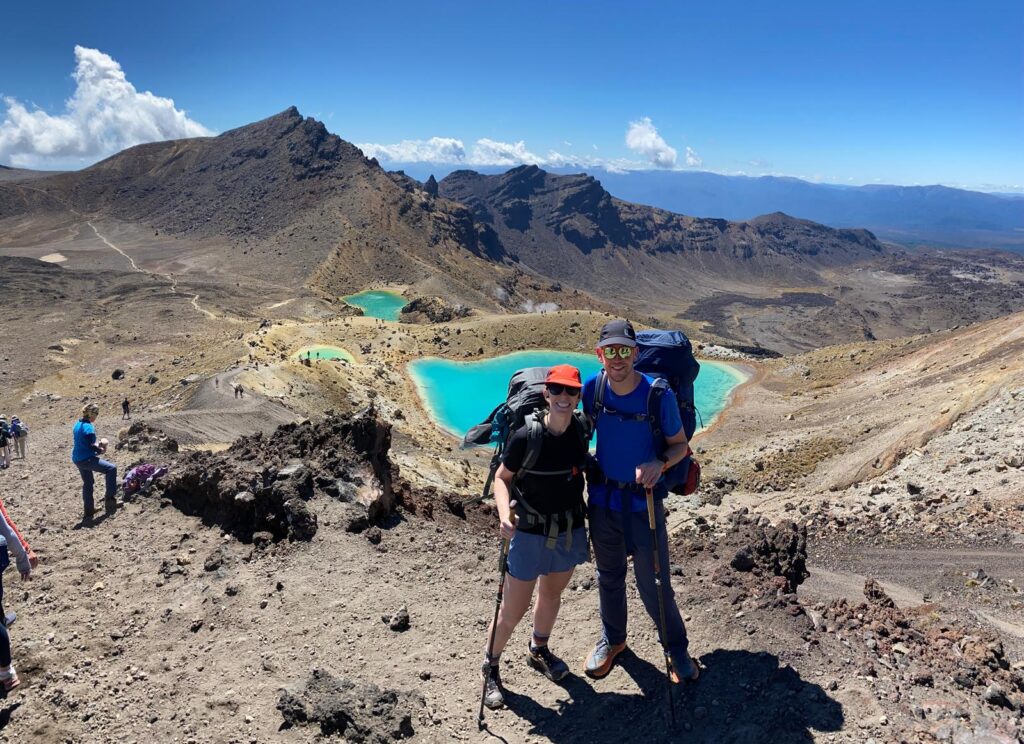 Two hikers pose on the Tongariro Alpine Crossing with the crystalline blue Emerald Lakes behind them in the summer time in New Zealand