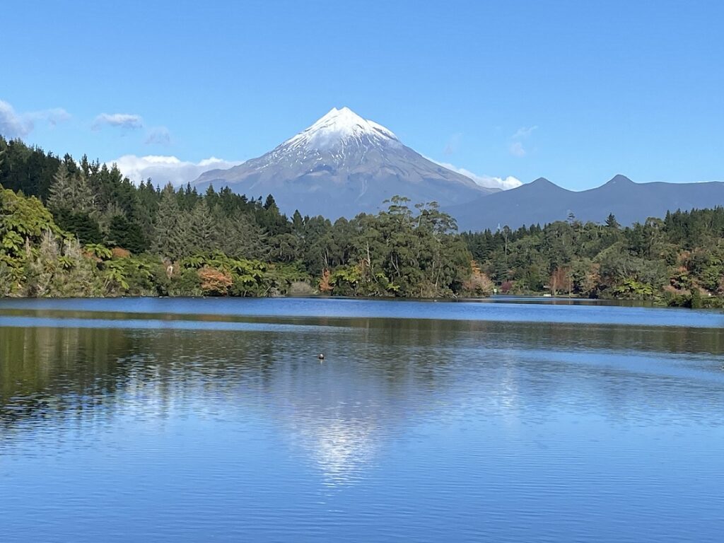 Mount Taranaki snow capped peak reflected in calm water of Lake Mangamahoe in foreground