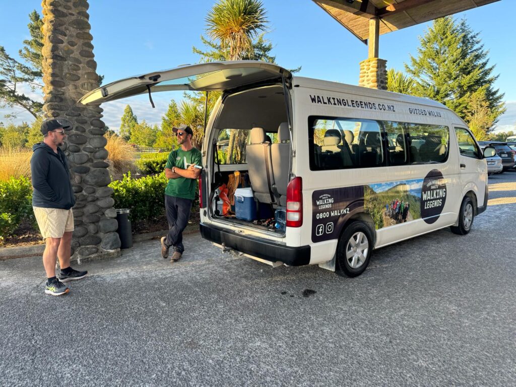 A hiking guide from Walking Legends rests casually against the tour vehicle parked outside The Park Hotel Ruapehu in discussion with guided walk guest about the Tongariro Alpine Crossing track
