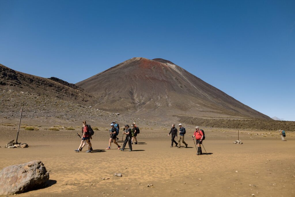 Hikers cross the flat brown earth of South Cirque on the Tongariro Alpine Crossing track with the peak of Mt Ngauruhoe in the background on a blue sky day