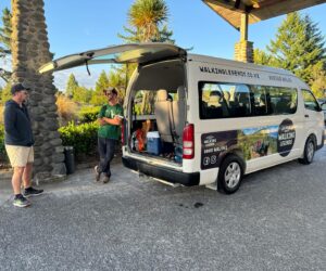 A hiking guide from Walking Legends rests casually against the tour vehicle parked outside The Park Hotel Ruapehu in discussion with guided walk guest about the Tongariro Alpine Crossing track on guided walks in Tongariro